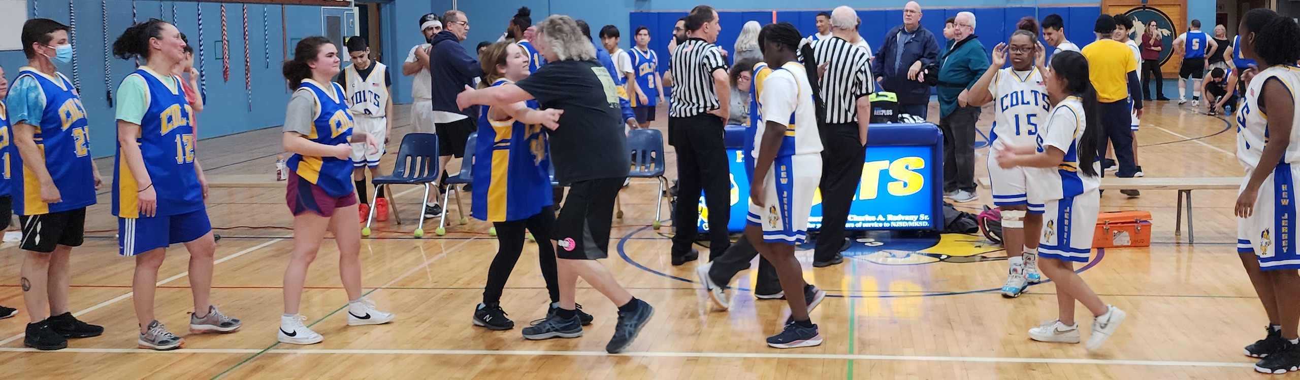 Alumni in Blue Colts Jersey in line to congratulate students in white Colts jersey at center court of MKSD gym. Various staff, students, and alumni, and two referees standing on court in background.